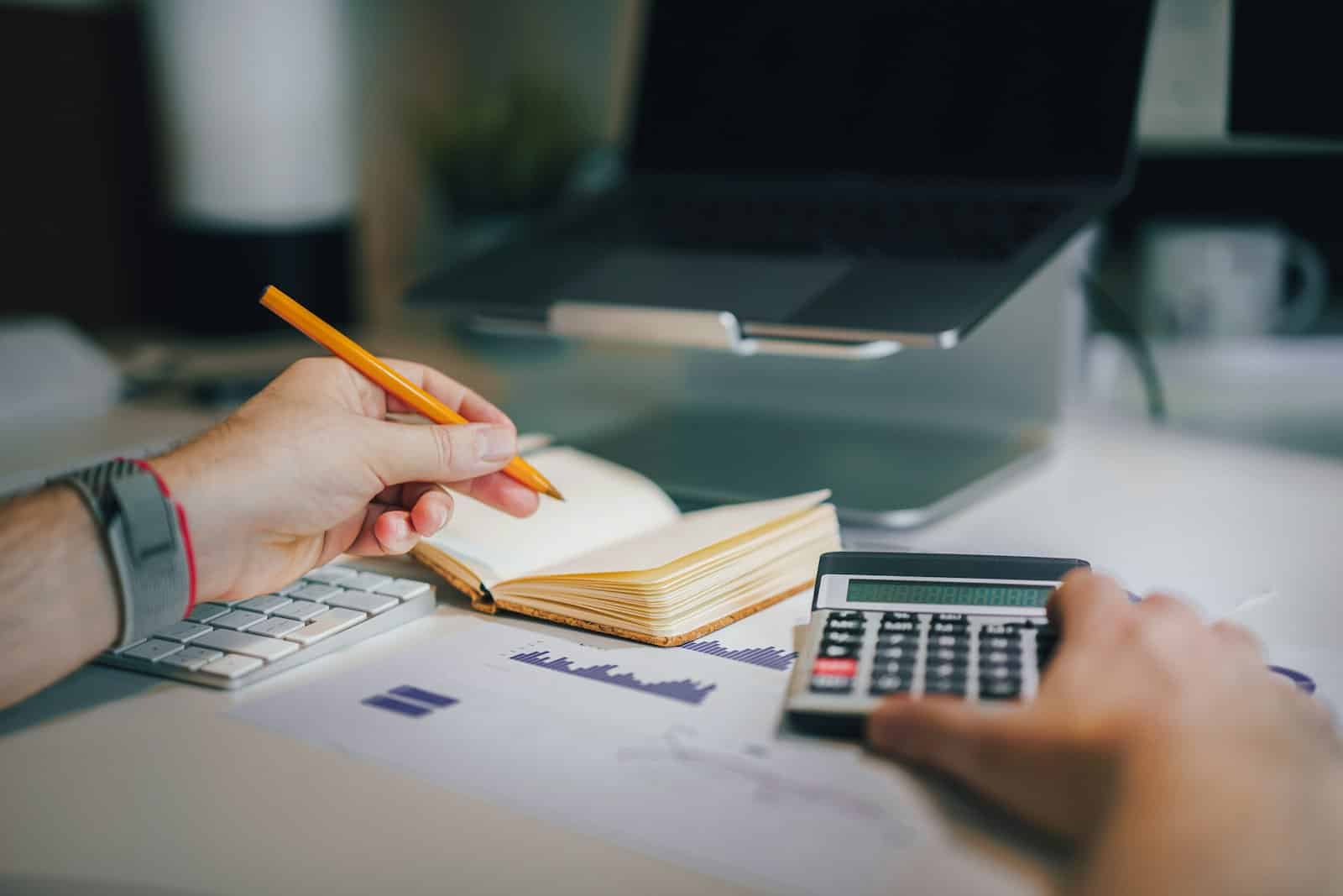 Une personne fait ses comptes avec une calculatrice et un carnet devant un ordinateur portable
